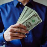 Close-up of a person in blue shirt holding multiple US dollar bills.