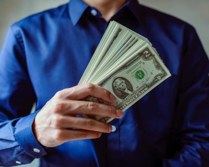 Close-up of a person in blue shirt holding multiple US dollar bills.