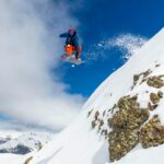 man in red jacket and blue pants sitting on snow covered mountain during daytime