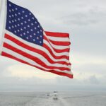 A vibrant American flag flutters on a boat with a vast ocean and cloudy sky backdrop.