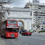 Iconic red double-decker bus passing Macedonia Gate in Skopje.