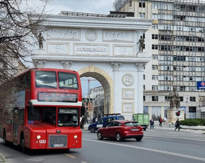 Iconic red double-decker bus passing Macedonia Gate in Skopje.