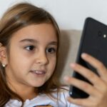 A young girl with long brown hair smiles while using a smartphone indoors.