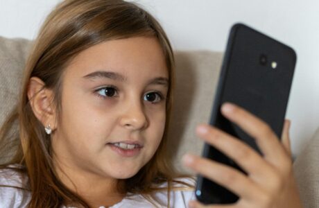 A young girl with long brown hair smiles while using a smartphone indoors.