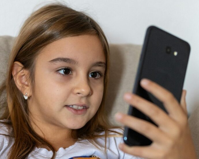 A young girl with long brown hair smiles while using a smartphone indoors.