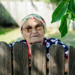 Portrait of an elderly woman in glasses and headscarf peeking over a wooden fence outdoors in Romania.