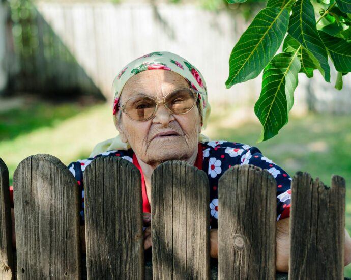 Portrait of an elderly woman in glasses and headscarf peeking over a wooden fence outdoors in Romania.