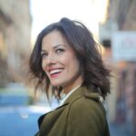 Portrait of a smiling woman standing on a city street in a green coat, looking away delightfully.