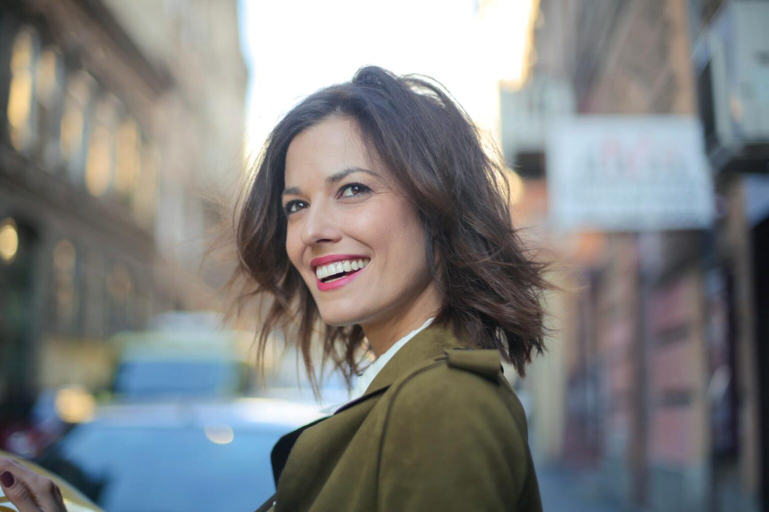 Portrait of a smiling woman standing on a city street in a green coat, looking away delightfully.