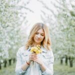 A young woman with blond hair holds dandelions in a blooming orchard, signifying springtime beauty.