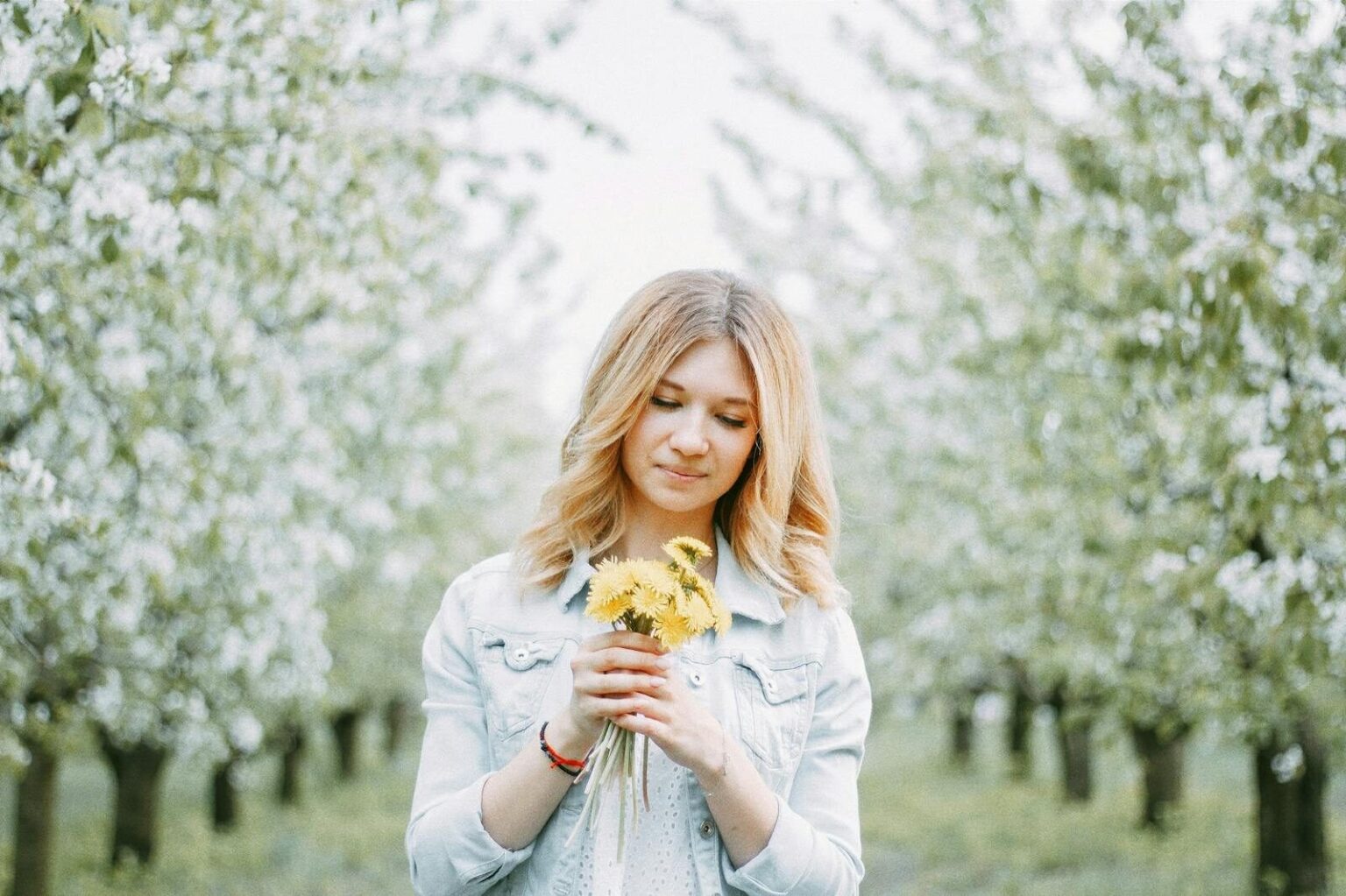 A young woman with blond hair holds dandelions in a blooming orchard, signifying springtime beauty.