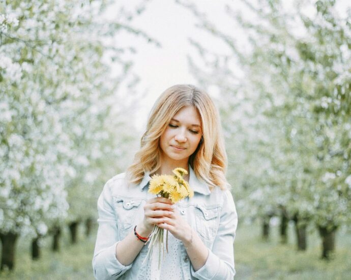 A young woman with blond hair holds dandelions in a blooming orchard, signifying springtime beauty.