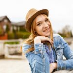 Smiling woman in denim and a hat enjoying a sunny outdoor day with a scenic background.