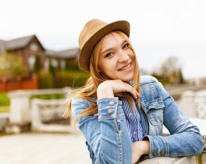 Smiling woman in denim and a hat enjoying a sunny outdoor day with a scenic background.