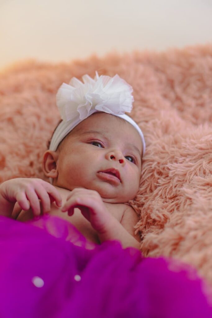 Charming portrait of a baby girl wearing a white hairband, lying on a fluffy pink blanket.