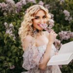 Blonde woman in a floral dress enjoying reading outdoors in a garden of lilac flowers.