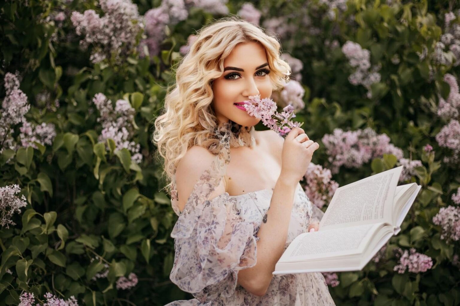 Blonde woman in a floral dress enjoying reading outdoors in a garden of lilac flowers.