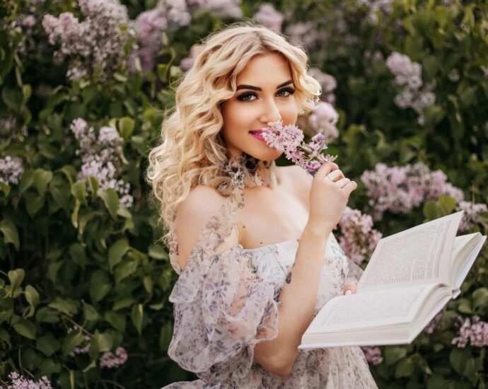 Blonde woman in a floral dress enjoying reading outdoors in a garden of lilac flowers.