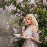 A fashionable woman in a sundress reads a book surrounded by blooming lilacs, exuding springtime elegance.