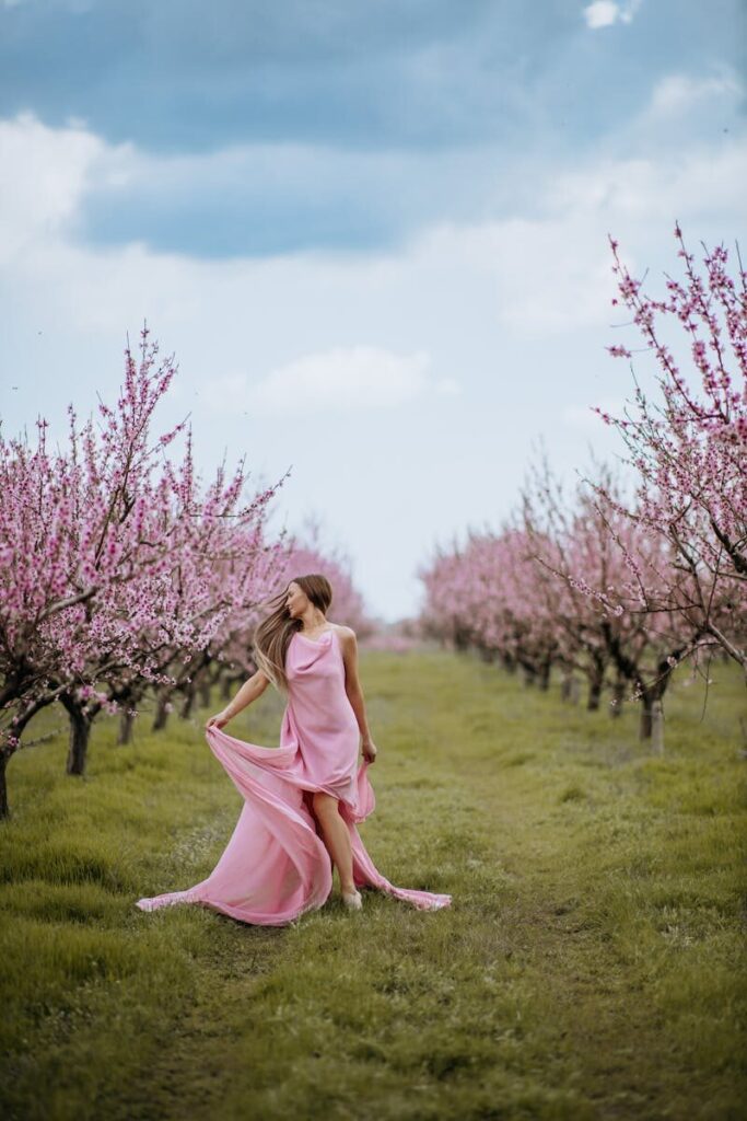 A woman in a flowing peach dress stands gracefully amidst a blooming orchard, evoking a sense of serene spring beauty.