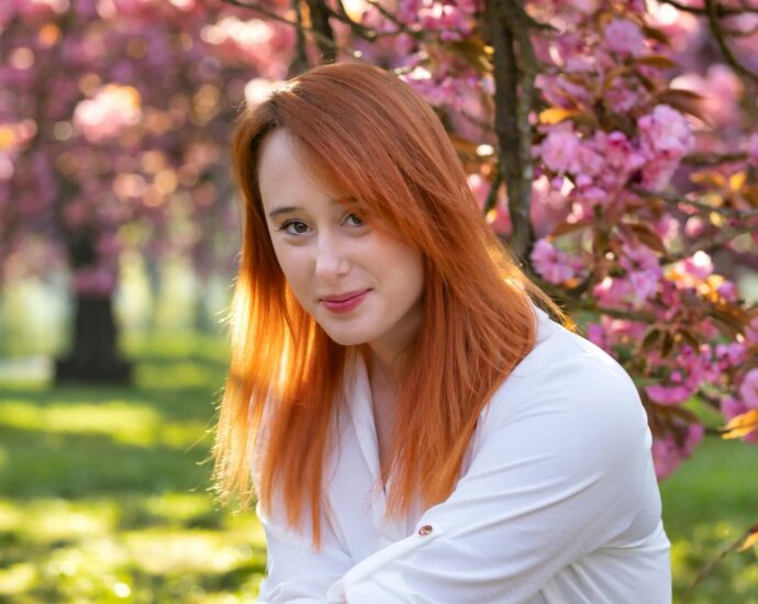 Portrait of a woman with red hair smiling among pink cherry blossoms in spring.