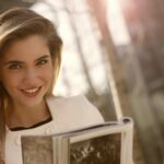 Smiling woman with long blond hair reading a magazine outdoors in sunlight.