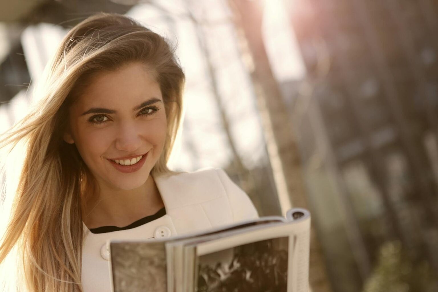 Smiling woman with long blond hair reading a magazine outdoors in sunlight.