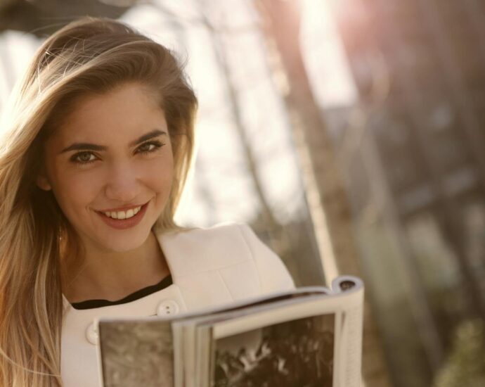 Smiling woman with long blond hair reading a magazine outdoors in sunlight.