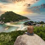 topless man sitting on cliff rock viewing Ko Nang Yuan isalnd, houses, and buildings under white and yellow sky