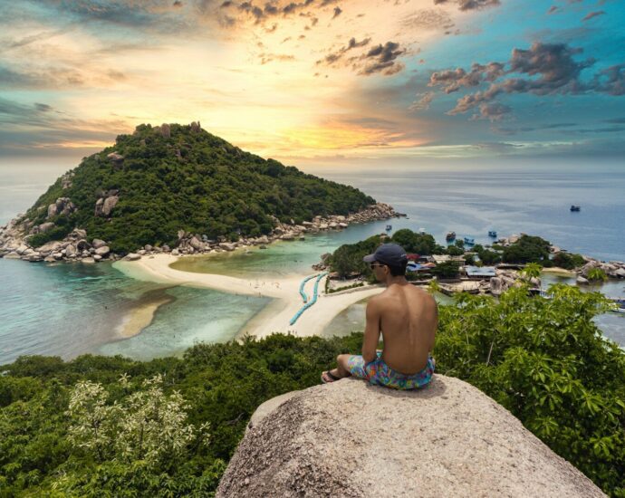 topless man sitting on cliff rock viewing Ko Nang Yuan isalnd, houses, and buildings under white and yellow sky