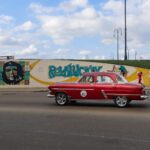 Vintage red car cruising by Cuban street art in Havana, featuring Che Guevara mural.