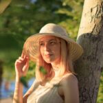 A woman in a hat enjoying a sunny day outdoors against a tree.