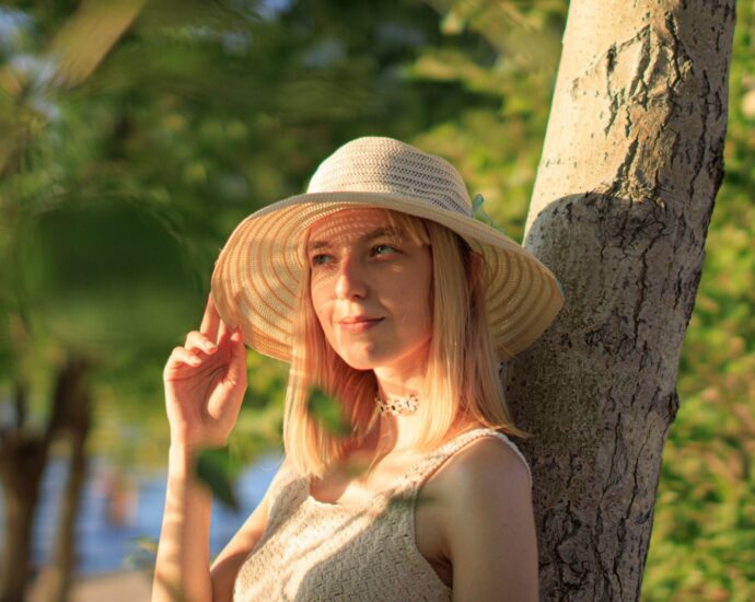 A woman in a hat enjoying a sunny day outdoors against a tree.