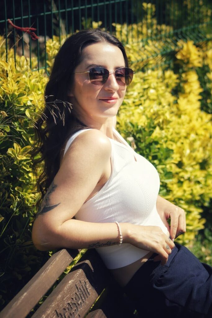 Stylish woman in white top and sunglasses relaxing on a park bench surrounded by lush greenery.