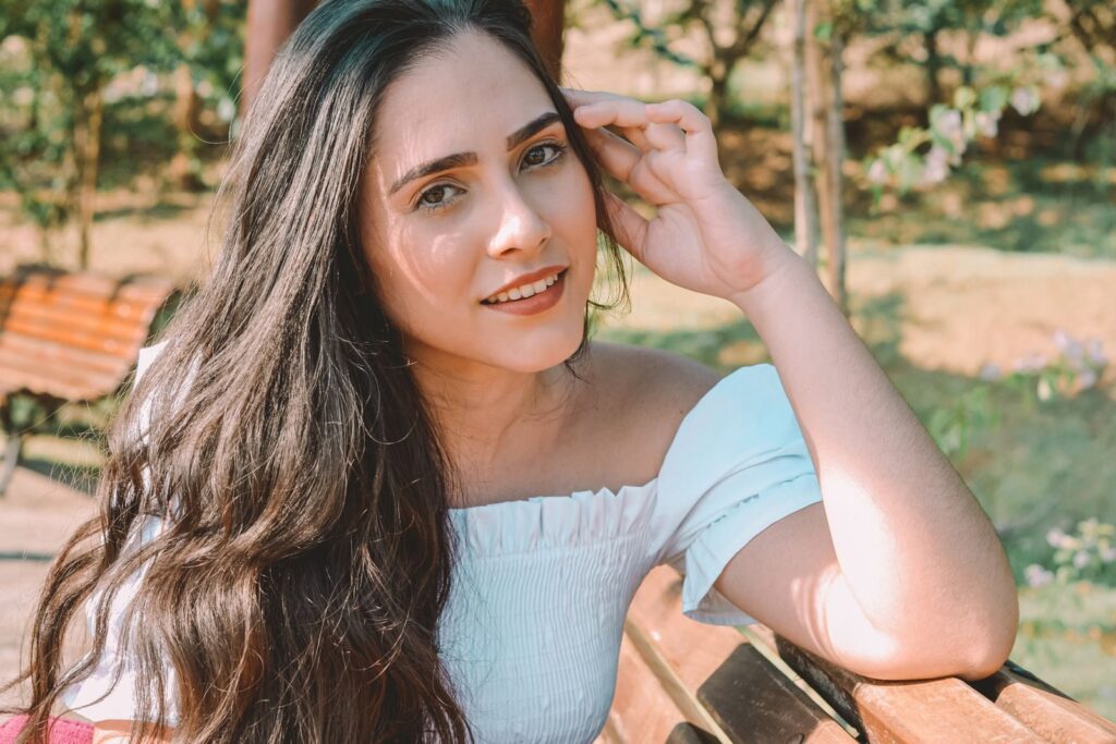 A young woman with long hair sitting on a bench in a sunny park setting.