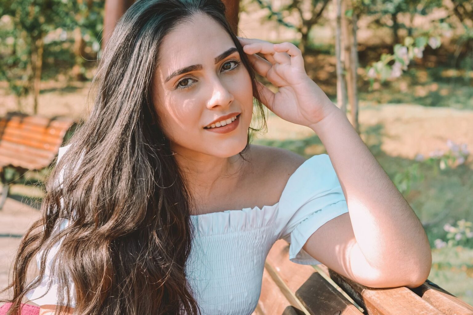 A young woman with long hair sitting on a bench in a sunny park setting.