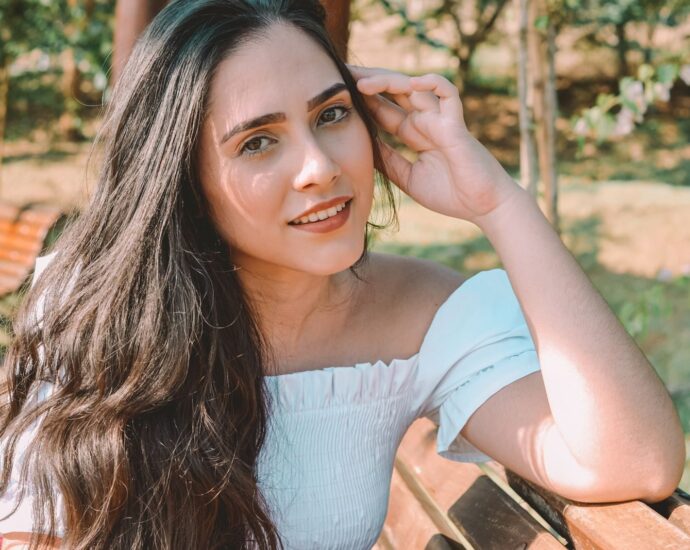 A young woman with long hair sitting on a bench in a sunny park setting.