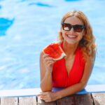 A cheerful woman in sunglasses enjoys watermelon by a vibrant pool.