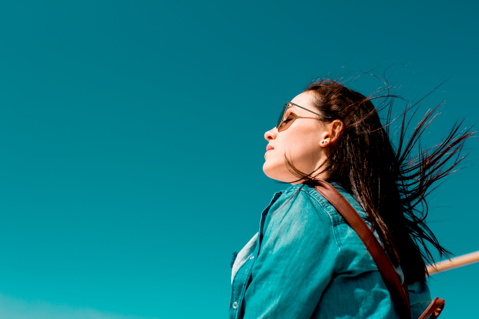 Fashionable woman outdoors in denim shirt with sunglasses under clear blue sky, enjoying sunny weather.