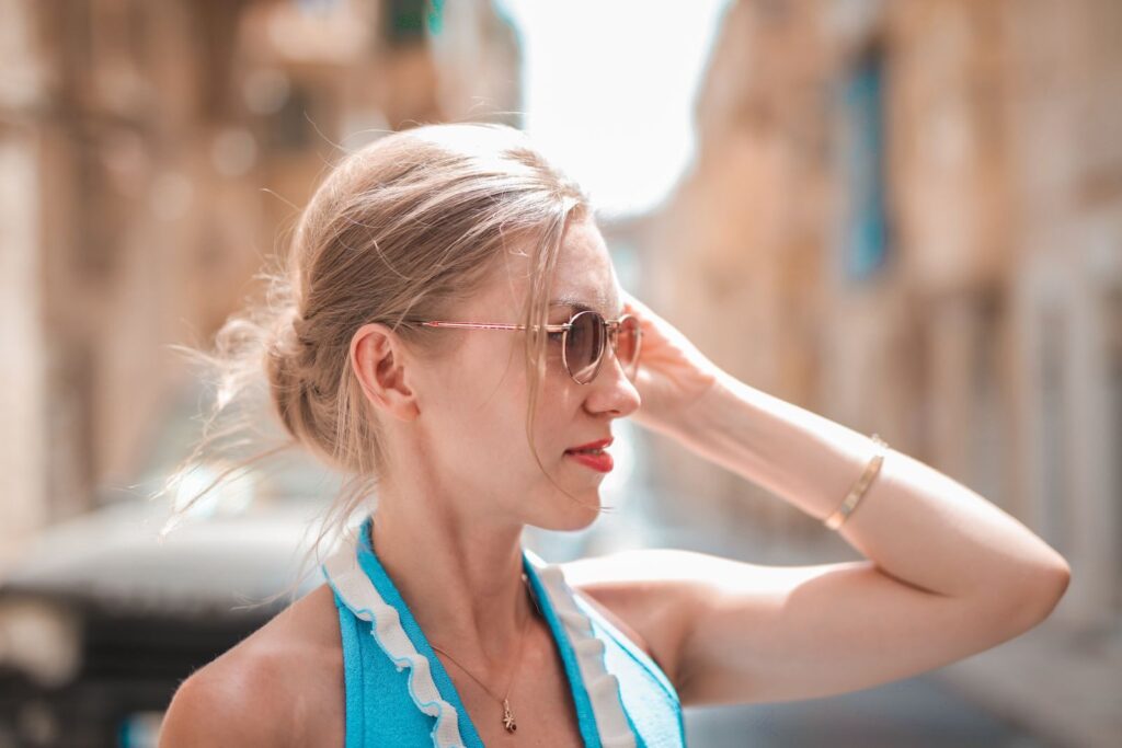 Elegant woman in sunglasses and blue dress stands on a sunlit street in Mgarr, Malta.