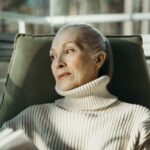 Elderly woman in a turtleneck sweater sitting comfortably indoors, looking pensive.