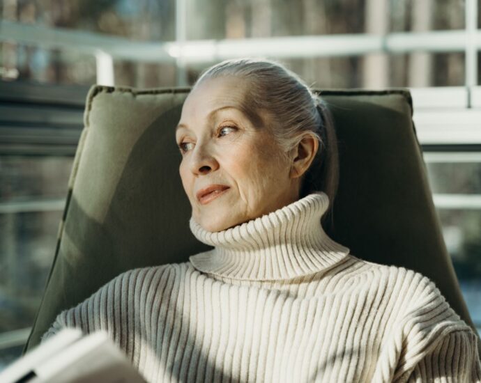 Elderly woman in a turtleneck sweater sitting comfortably indoors, looking pensive.