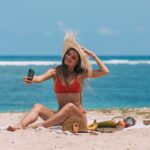 Woman in bikini taking a selfie on a sunny beach with ocean backdrop.