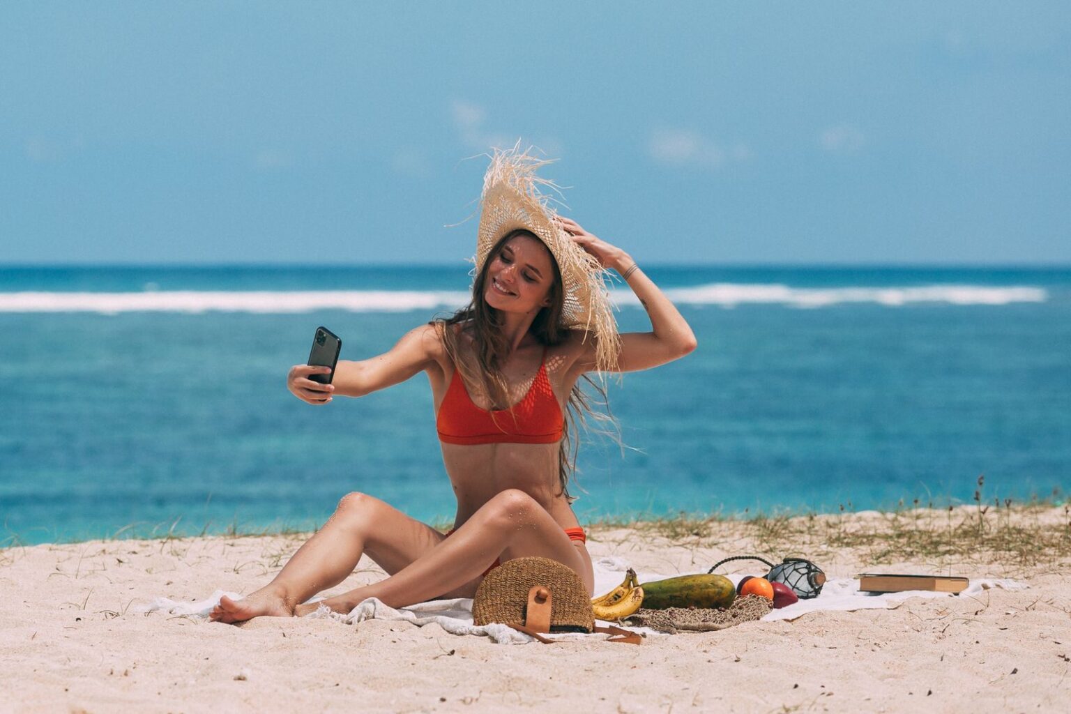 Woman in bikini taking a selfie on a sunny beach with ocean backdrop.