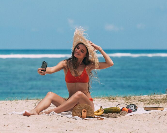 Woman in bikini taking a selfie on a sunny beach with ocean backdrop.