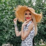 Charming blonde woman in a straw hat smiling outdoors in a floral sundress.