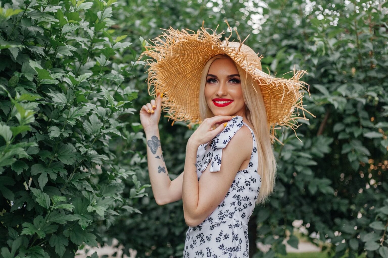 Charming blonde woman in a straw hat smiling outdoors in a floral sundress.