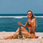 Woman in bikini relaxing on a sunny beach with tropical fruits and a refreshing drink.