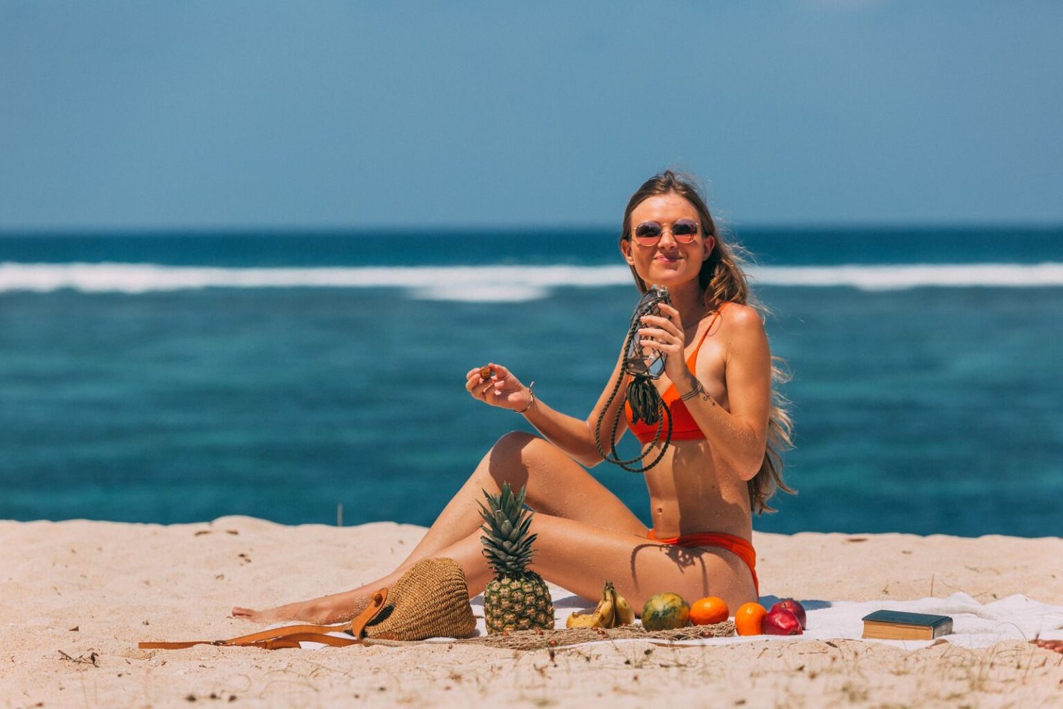 Woman in bikini relaxing on a sunny beach with tropical fruits and a refreshing drink.