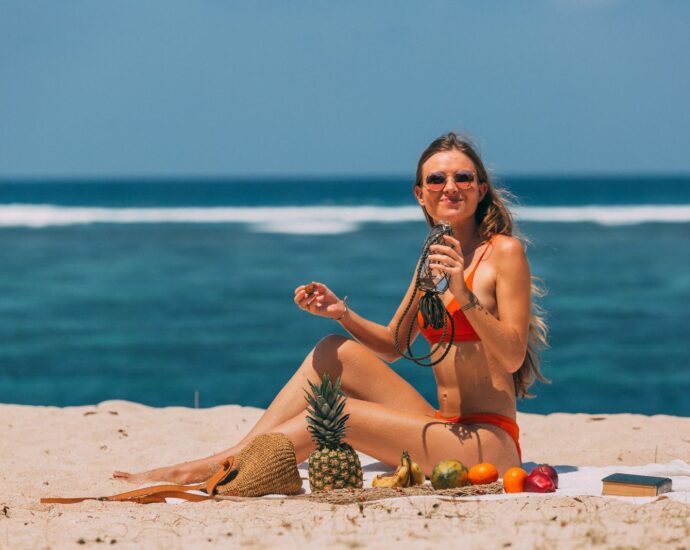 Woman in bikini relaxing on a sunny beach with tropical fruits and a refreshing drink.